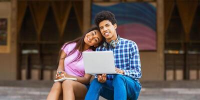 un adolescente negro Pareja sentar en un agacharse afuera, un ordenador portátil en el adolescente mans regazo mientras él sonrisas a el cámara. su brazo es alrededor su novia, quien es mirando hacia el cámara y sonriente foto
