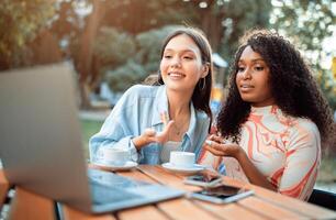 Two women are seated at a table in a cafe setting. One woman is looking at the laptop and speaking, while the other woman listens intently. Both women have coffee cups in front of them. photo