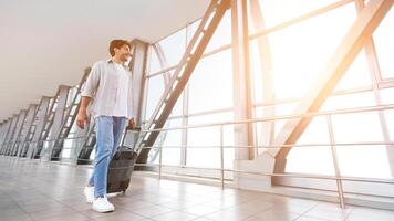 Enjoying vacation. Cheerful man walking with luggage in airport terminal, looking at sunset in window, empty space photo