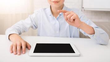 A young boy sits at a white desk in a bright room. He is wearing a light blue button-down shirt and is using a digital tablet with a black screen. He is pointing with his index finger at the tablet. photo