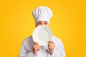 Funny Chef Guy Peeking Out Of Plate Looking At Camera Standing In Studio Over Yellow Background. Panorama photo