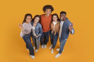 A group of five friends, all dressed in casual attire, pose together on a bright yellow background. They look happy and excited, with their arms around each other, view above photo