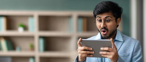 Indian man wearing glasses expresses surprise while using a tablet in a contemporary office environment. The scene features shelves filled with books and decor in the background. photo