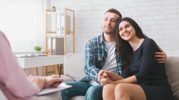A couple sits on a couch in a therapy session. They are holding hands and looking off to the side, as if in deep thought or reflection, seem to be in a loving and supportive relationship photo