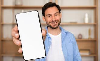 A man stands in a bright, inviting room, smiling as he holds out a blank smartphone. The background features shelf decor, creating a warm atmosphere photo