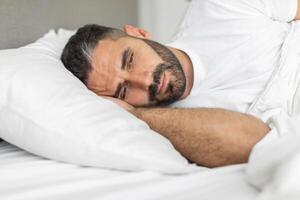 A man is lying in bed with his arm under his head. He is resting his head on a white pillow, and he has a small beard. The man appears to be sleepless, suffering from depression photo