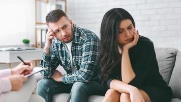 A couple sits on a grey couch in a therapy session, looking down with expressions of sadness or worry. A therapists hand is visible in the foreground, holding a pen and notepad photo