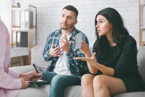 A young couple sits on a couch during a therapy session. They are discussing their smartphone usage, with the woman holding a smartphone in her hand and pointing to it with her left hand photo