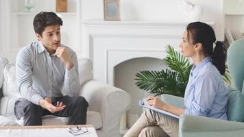 A man is seated on a light-colored couch, holding his chin thoughtfully while engaging in a counseling session with a female therapist, therapist is taking notes on a clipboard and appears attentive photo