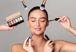 A woman with eyes closed is having various makeup products applied on her face by multiple hands around her. She holds a serene expression as brushes and palettes are used to enhance her makeup look. photo