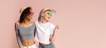 Two young women are standing in front of a solid pink backdrop. Both women are wearing sunglasses and smiling. The woman on the right is pointing to the left of the frame, copy space photo