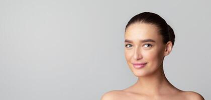 A young woman is smiling while standing in front of a plain gray background. Her expression is cheerful and relaxed as she looks directly at the camera, panorama with copy space photo