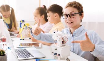 A young boy sits in a classroom setting with other students. He is smiling and pointing at a small robot he has built, demonstrating his learning and enthusiasm for STEM education. photo