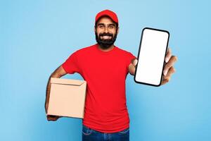 Indian delivery man wearing a red shirt and hat is holding brown cardboard box in his left hand and smartphone with blank screen in his right hand. He is smiling and looking directly at the camera. photo