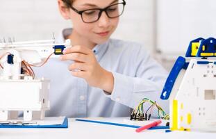 A young boy wearing glasses is working on a robotics project. He is holding a small, blue, electronic component, while a white robot with wires and a circuit board is on the table in front of him. photo