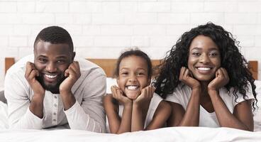 Portrait of happy black family lying on bed together, propping chin up with hands and smiling to camera, copy space photo