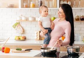 A woman standing in a kitchen holding a baby in her arms. The woman appears to be cooking or preparing food while caring for son. The kitchen is well-lit and appears to be a typical household setting. photo