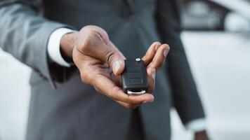 Unrecognizable man in a gray suit holds out a car key, showing the silver key fob with multiple buttons. The man is standing in front of a white car, with the cars exterior out of focus. photo