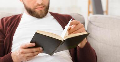A man is seated on a couch, engrossed in reading a book. The man appears focused and relaxed, surrounded by a quiet atmosphere, cropped photo