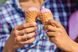 A close-up shot of two hands holding pink ice cream cones. The ice cream is melting slightly and the waffle cones are crisp. The hands are in focus, while the background is blurred photo
