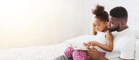A little Black girl sits on her fathers lap as they read a book together in bed. The father is smiling and looking at his daughter, who is pointing at the book. The scene is warm and loving photo