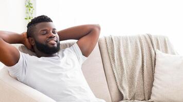 This image shows a black man with a beard relaxing on a white couch. He has his arms behind his head and his eyes are closed as he leans back into the cushions, copy space photo