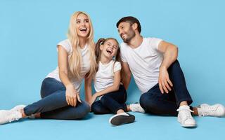 A family of three, a man, a woman, and a young girl, are sitting on a blue backdrop. The family is laughing and looking happy, with the girl in the middle smiling wide. photo