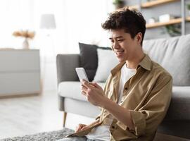 Testing New App. Handsome Asian Man Using Smartphone While Relaxing Leaning On Couch At Home, Happy Guy Sitting On Floor In Living Room, Browsing Internet Or Messaging With Friends, Free Copy Space photo