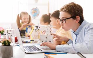 A teenage boy wearing glasses sits at a desk in a classroom, using a laptop computer. He is surrounded by other students, including a girl who appears to be working on a small robot. photo