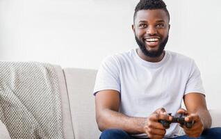 Black young man with a beard sits on a couch while playing a game. He is smiling and holding a game controller. The background is a simple white wall, copy space photo
