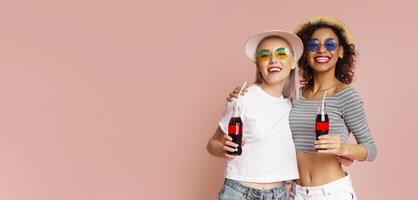 This image features two women in summer hats and sunglasses, smiling and holding soda bottles against a pink background. They are both wearing casual clothing, copy space photo