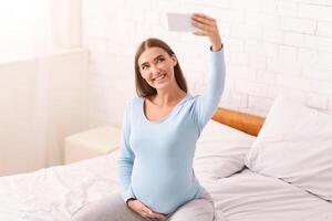 Happy Pregnancy Selfie. Smiling Pregnant Girl With Smartphone Making Self-Portrait Sitting On Bed At Home. Selective Focus photo