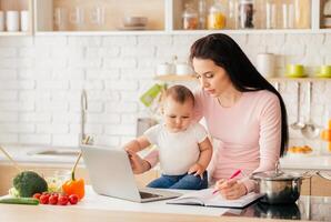 A mother multitasks in a bright, modern kitchen, working on her laptop and taking notes while holding her baby. Fresh vegetables and a pot on the stove are visible in the background. photo