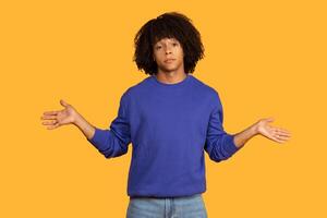 A young man with an curly hairstyle is standing in front of a yellow background. He is wearing a blue sweater and jeans. He is gesturing with both hands, palms facing outwards photo