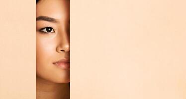 Asian woman face peeking from behind a piece of beige paper. Only her eye and part of her face are visible, creating a sense of mystery and intrigue, copy space photo