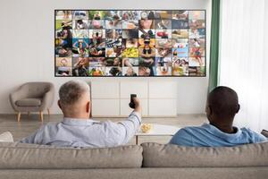 Two men are seated on a couch in a living room, watching television. The television is displaying a grid of images, and the man on the left is holding a remote control. photo