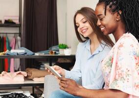 Two multiethnic women are seated on a couch, both looking intently at digital tablet held between them, visiting fashion store, checking new collection photo
