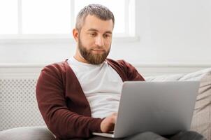 A man is seated on a couch, focusing intently on his laptop screen. His hands are busy typing on the keyboard as he works or browses online. photo