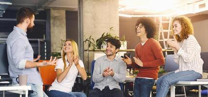 A casually dressed diverse group of colleagues is gathered in a brightly lit modern office, clapping and smiling, showing support and appreciation during an engaging team meeting photo