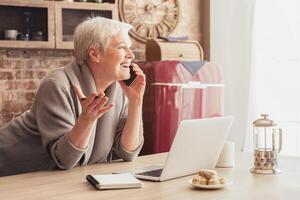 Elderly woman with short, gray hair sits at a kitchen table, talking on her phone. She is smiling and gesturing with her free hand. A laptop is open in front of her and a notebook is lying next to it. photo