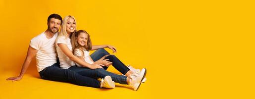 A joyful family of three sits closely together on the floor, dressed in matching white t-shirts and casual jeans. The vibrant yellow backdrop enhances the warmth of their smiles, copy space photo
