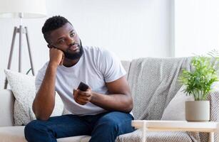Black man sits on a couch in a living room, looking bored. He is holding a remote control in his hand and has a thoughtful expression on his face. The room is decorated with a houseplant photo