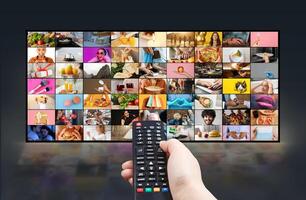 A persons hand holds a remote control and points it towards a large flat screen television. The TV screen displays a variety of colorful, diverse content in a grid format photo