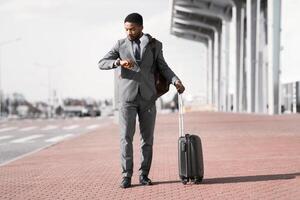 An African American man with luggage is checking the time near an airport entrance. The atmosphere is dynamic and contemporary, reflecting a busy travel schedule. photo