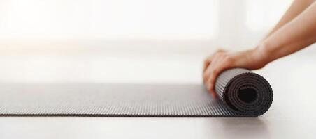 A person is shown rolling up a yoga mat on the floor. The individuals hands are carefully folding and tucking the mat to ensure a neat and compact roll, cropped, panorama photo