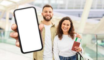 A beaming couple stands in an airport terminal, the man holding out a smartphone with a blank screen towards the camera, while the woman beside him clutches a passport and boarding pass. photo