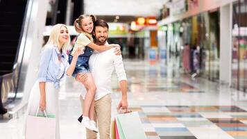 A smiling father carries his daughter on his shoulders as they walk through a modern shopping mall with their mother, who is carrying a shopping bag, panorama with copy space photo