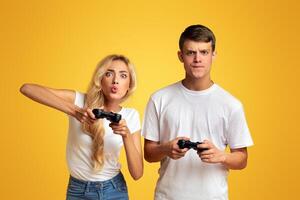 Young guy and girl emotionally playing game with joysticks, grimacing in excitement over yellow background. Panorama photo