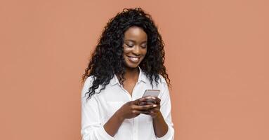 A young black woman with long curly black hair is smiling while looking at a smartphone in her hands. She is wearing a white button-up shirt and is standing in front of a peach-colored background. photo
