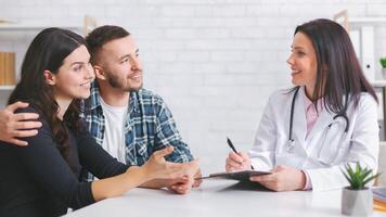 European couple sits at a table with woman doctor in a white coat. The couple is engaged in a conversation, listening intently as the doctor takes notes photo